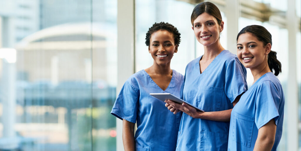 3 smiling nurses in front of building, woman in the middle is holding a tablet